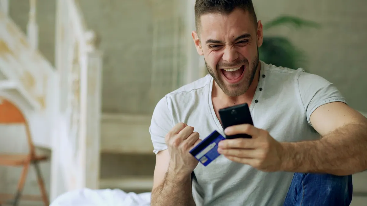 Hombre joven celebrando con el puño en alto mientras mira su teléfono y una tarjeta